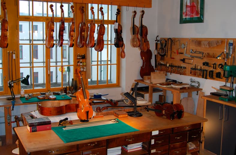 Werkbank im Geigenbauatelier mit hängenden Violinen und Cello vor Sprossenfenstern